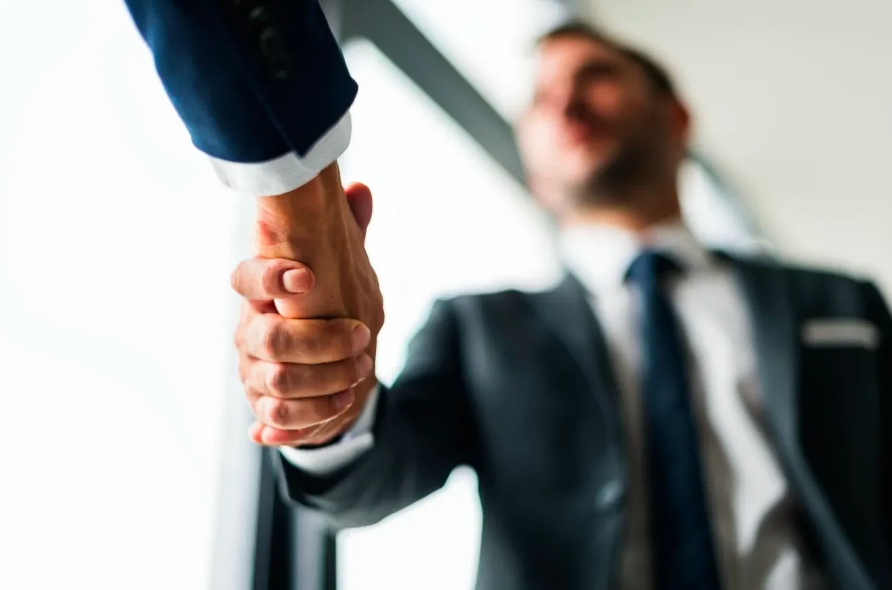 Two men in business suits shaking hands, with the smiling man in a tie blurred in the background