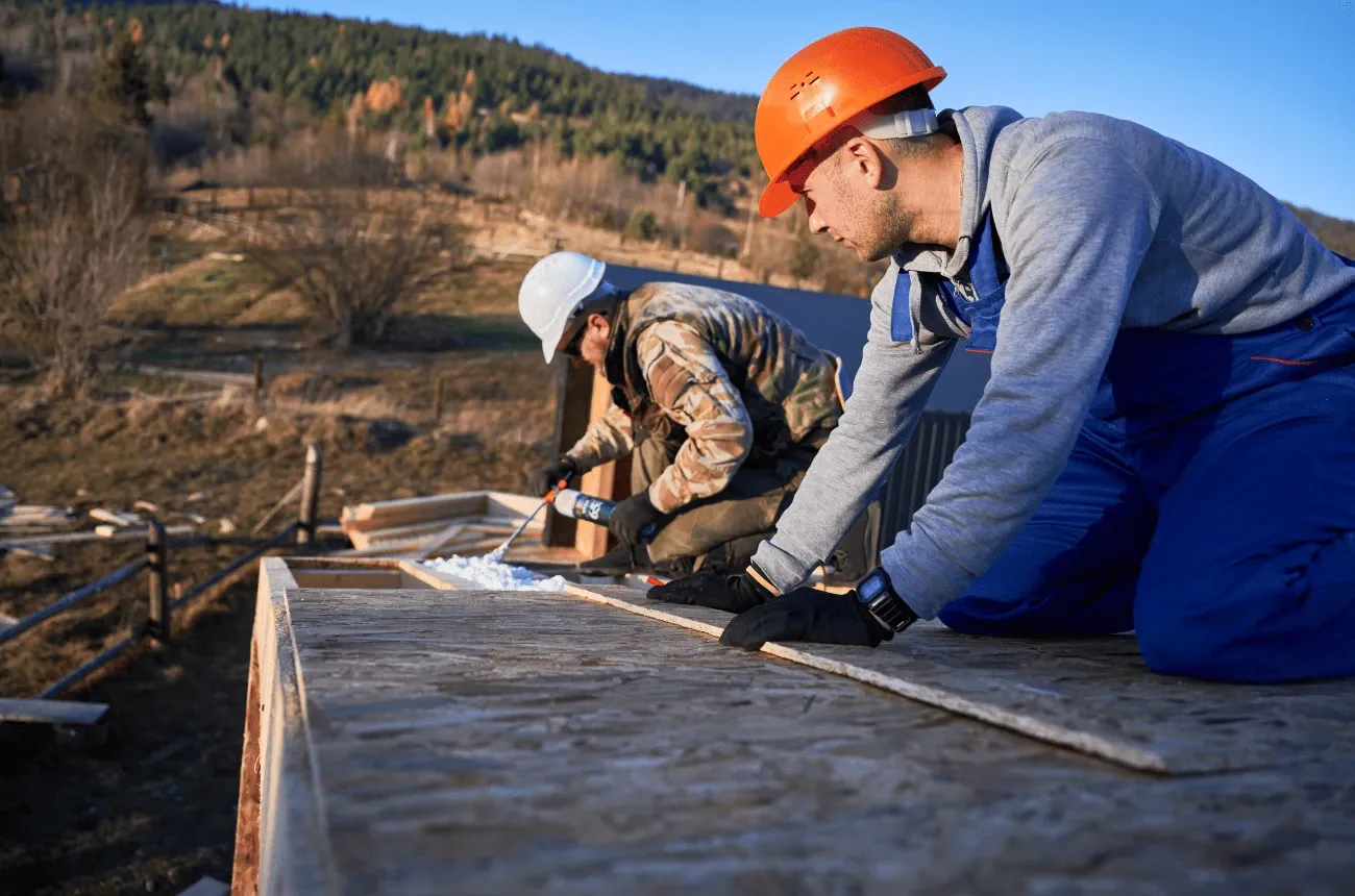 Two builders in hard hats kneel on a wooden roof frame, one applying spray foam, with hills in the background