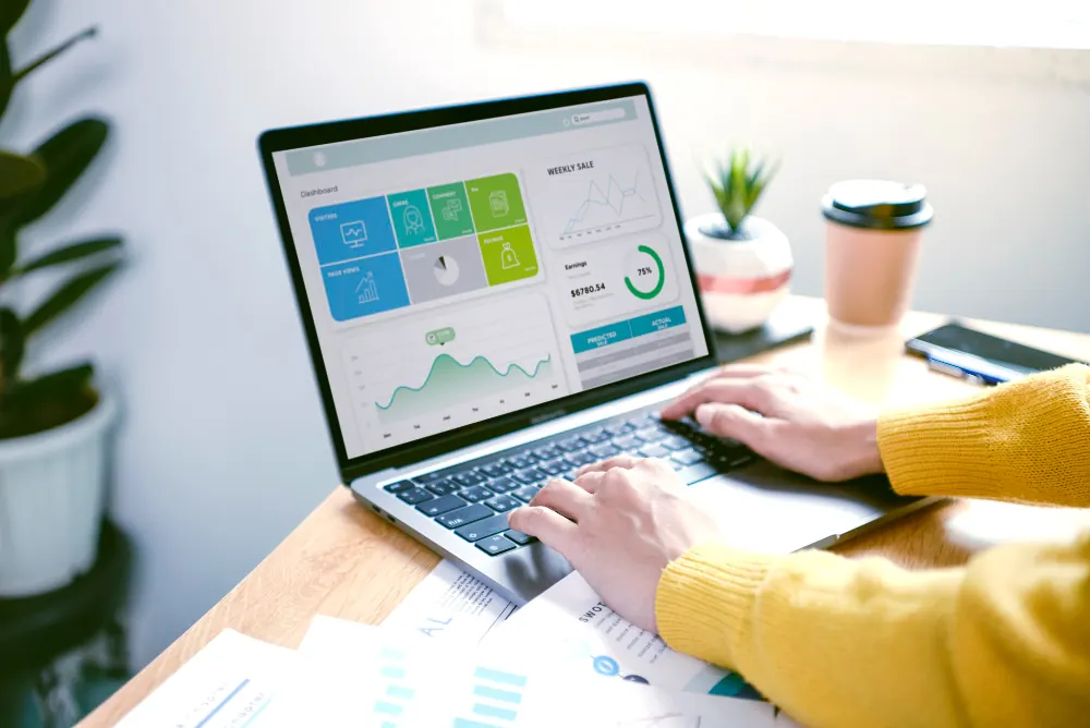A person in a yellow sweater typing on a laptop that shows a sales dashboard with charts